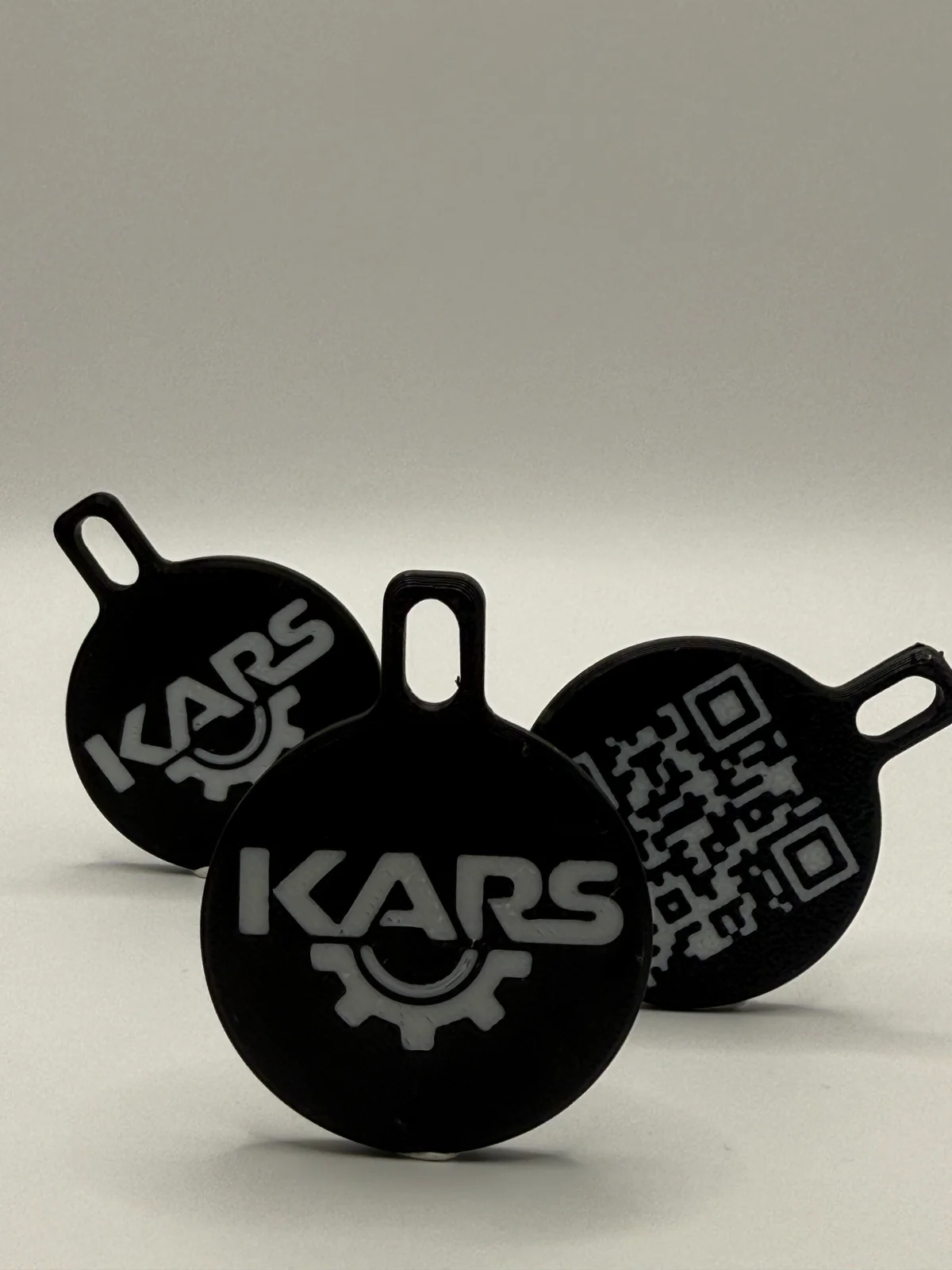 Trois porte-clés Kars noirs et blancs, avec le logo visible sur deux faces et un QR code sur la troisième.